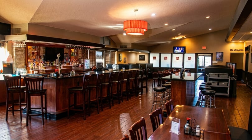 A spacious restaurant interior with a long wooden bar, high stools, and dining tables.