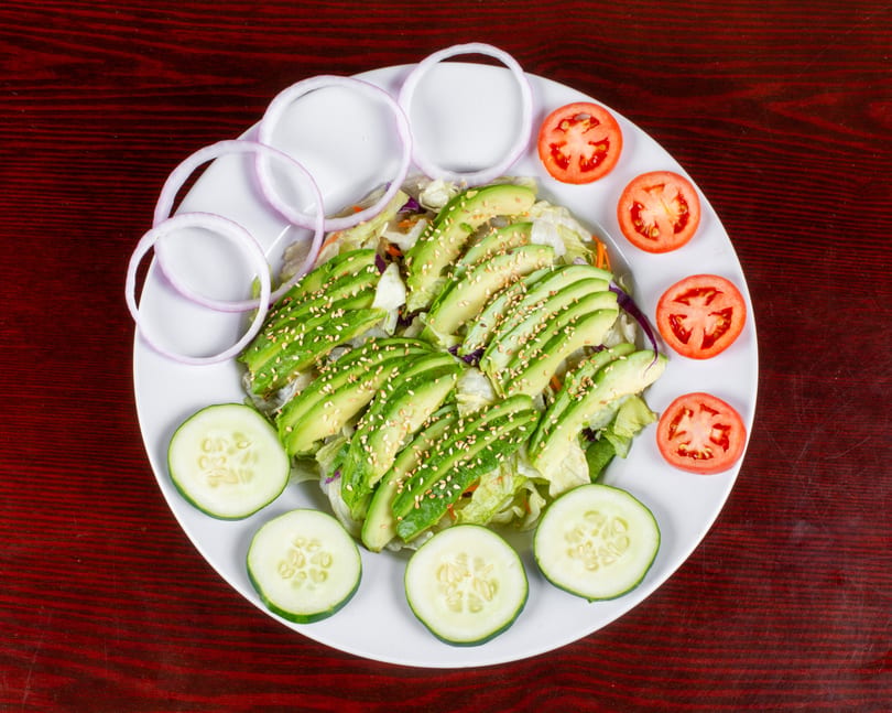 Avocado salad with lettuce, sesame seeds, red onion, tomato, and cucumber slices on a white plate.