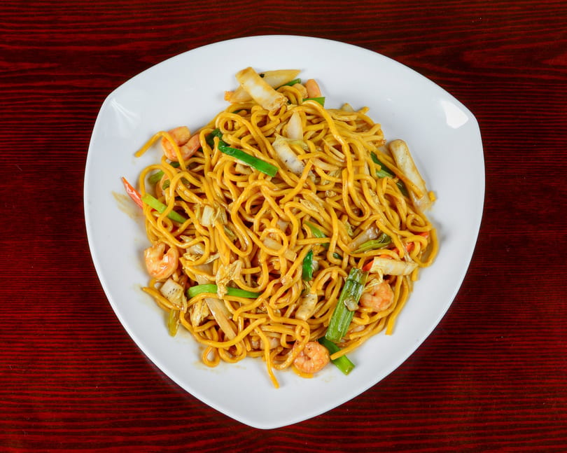 A white plate filled with golden noodles, shrimp, and mixed vegetables on a dark red wooden table.