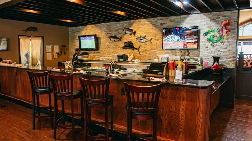 Sushi bar with wooden stools, stone wall, fish decorations, and two TVs showing sports.